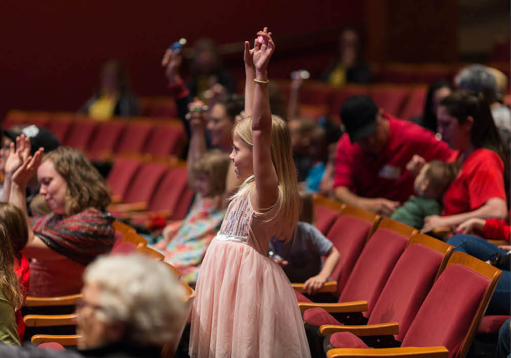 Girl standing in the audience with her arms raised above her head